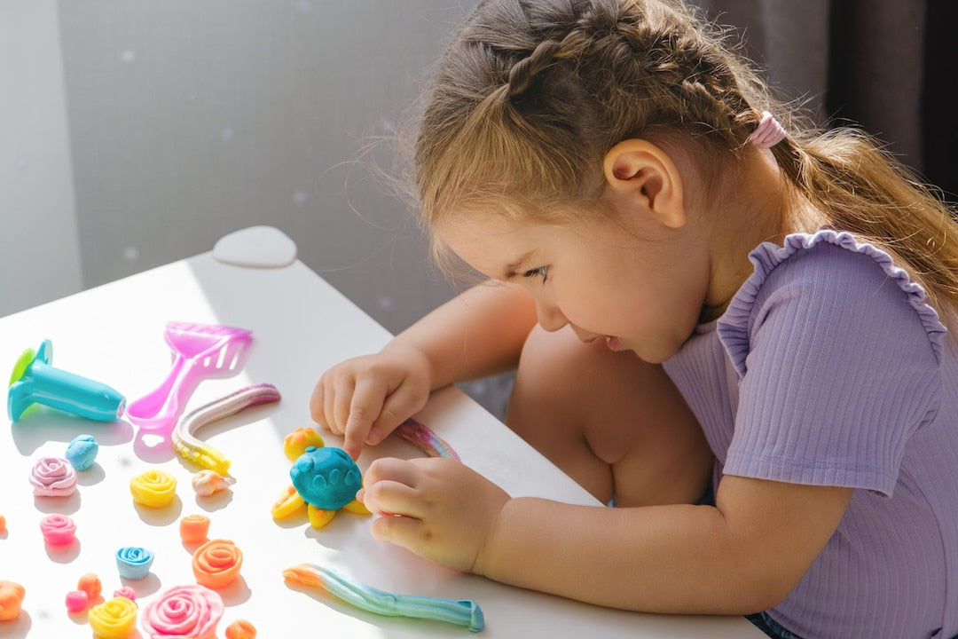 A little girl playing calmly with colorful air dry clay from OLIHUG creative kit on a sunny table. Montessori-inspired sensory play encourages focus, imagination, and relaxation without screens. Perfect for kids and parents to create, connect, and enjoy peaceful moments together. OLIHUG – Create Wonder  Peace.