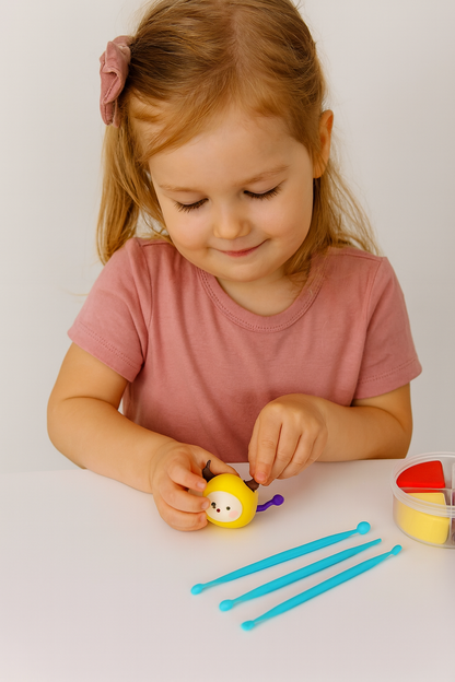 A young girl wearing a soft pink sweater is sitting at a light wooden table, shaping colorful air-dry clay into cute zodiac-themed figurines. Her small hands gently mold the clay tools and bright pastel materials, creating adorable animal shapes. The natural daylight and neutral background highlight the creative, hands-on process, making it a perfect depiction of mindful, screen-free playtime for kids and families.
