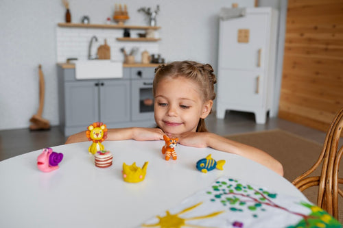 Smiling young girl sitting at a table, playing with colorful animal-shaped clay figures in a cozy home environment. A joyful example of creative, screen-free activity that supports focus, imagination, and child development.
